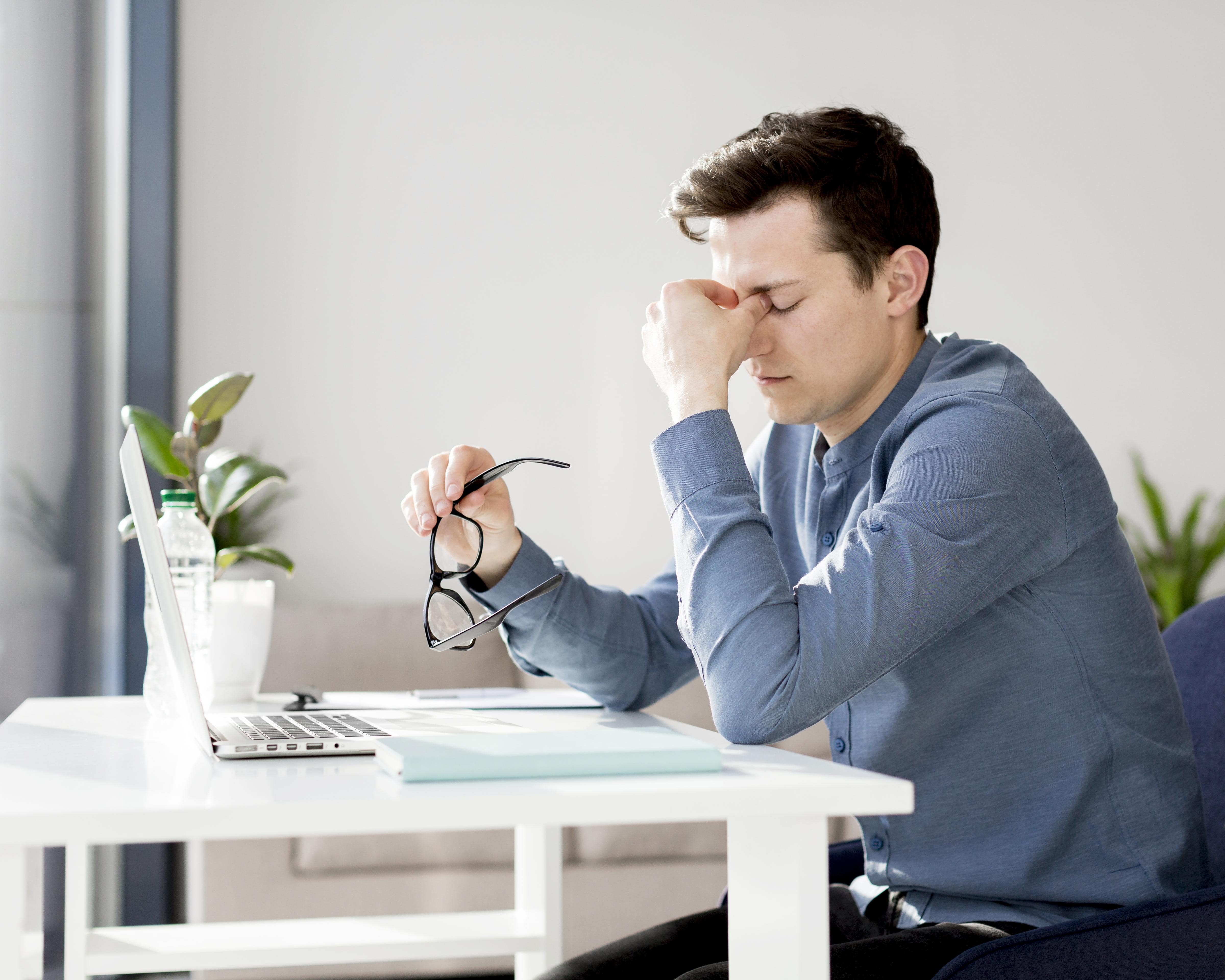 Homem muito frustado no escritório, com óculos na mão e pressionando a testa com os dedos