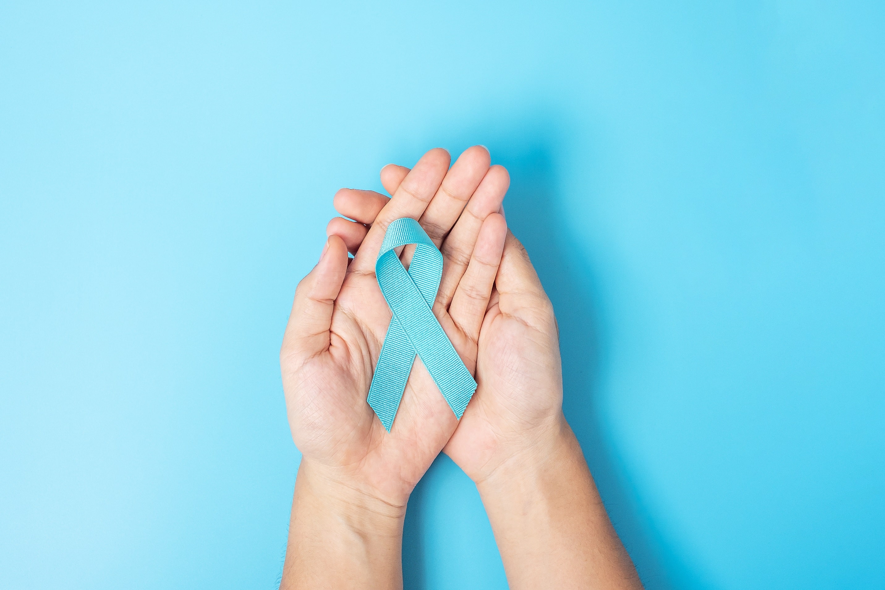 mãos masculinas segurando o laço azul, simbolo da campanha contra o câncer de próstata, com um fundo azul